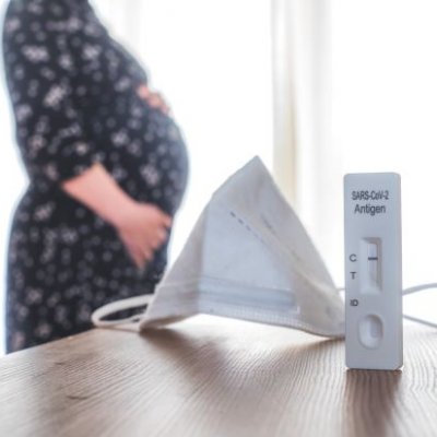 A medical mask and COVID-19 test on desk with pregnant woman in background. Adobe